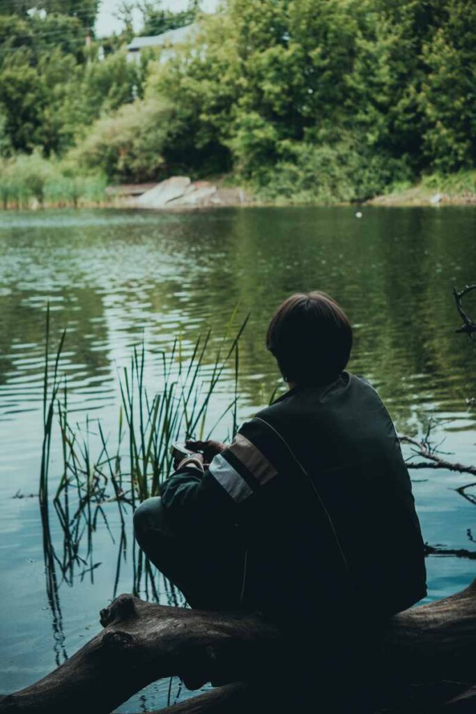 Person sitting by a calm lake surrounded by green trees, contemplating mental clarity during water fasting