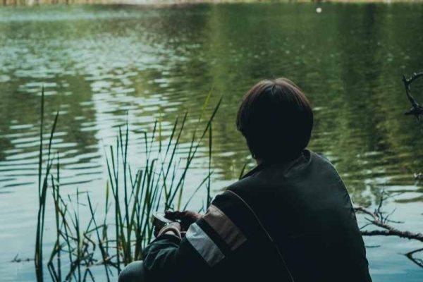 Person sitting by a calm lake surrounded by green trees, contemplating mental clarity during water fasting
